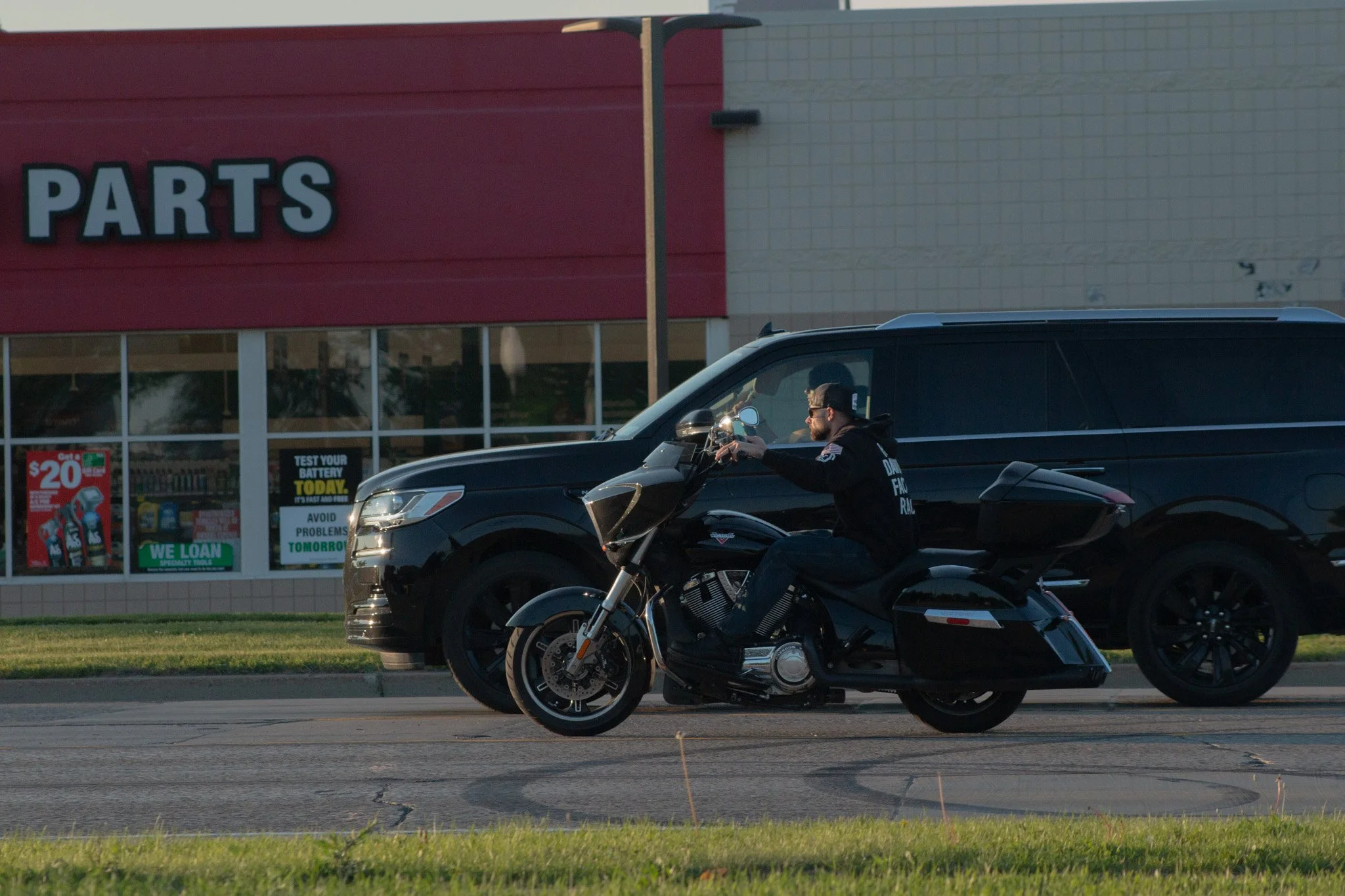A man in a black hoodie riding a black motorcycle on a city street in front of a parts store