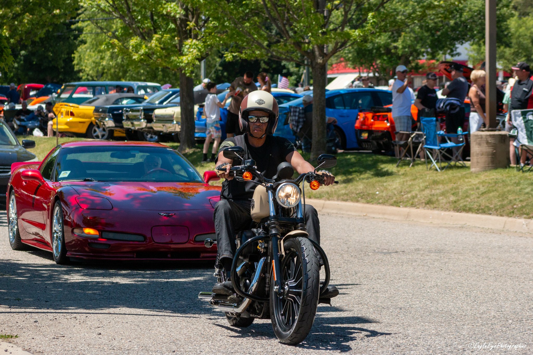 A man wearing a helmet and sunglasses riding a motorcycle past a red sports car during a car show, with several vintage and modern cars and people gathered in the background under green trees.