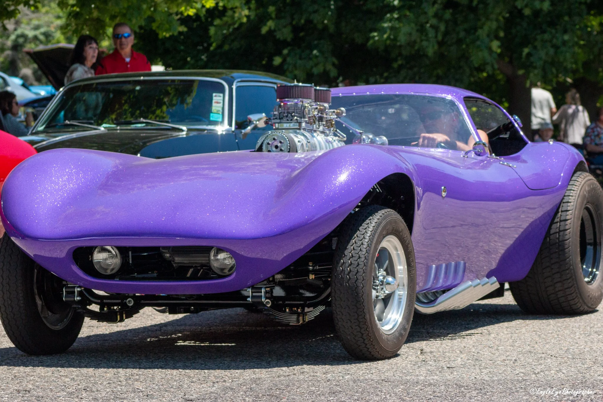 A purple classic car with a prominent engine modification displayed at an outdoor car show, with people and trees in the background.