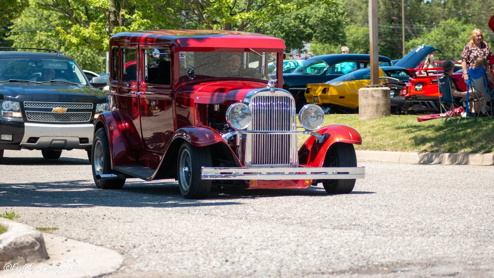 A vintage red automobile parked at a car show with other classic and modern vehicles in the background, and people sitting on lawn chairs under trees.