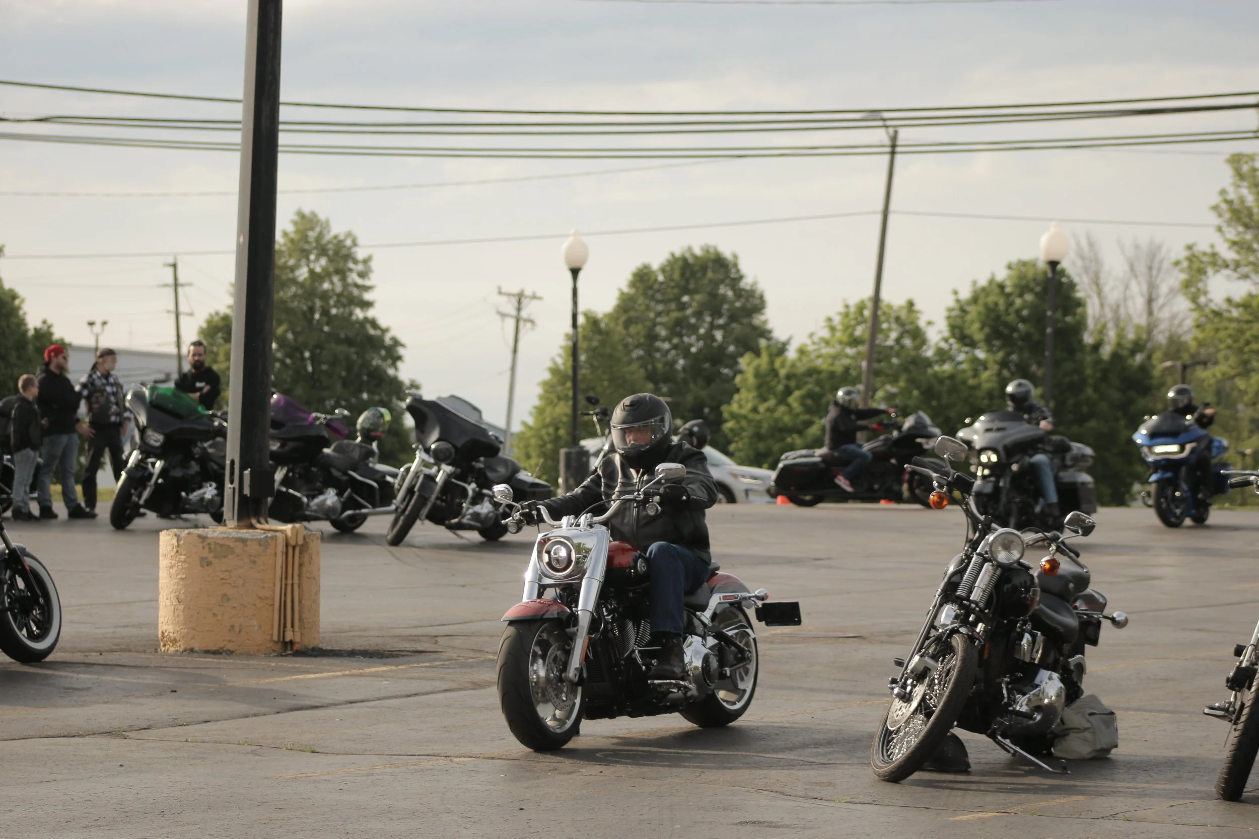 Motorcyclists riding bikes in a parking lot with other bikes parked and a few people standing nearby, trees, and power lines in the background.