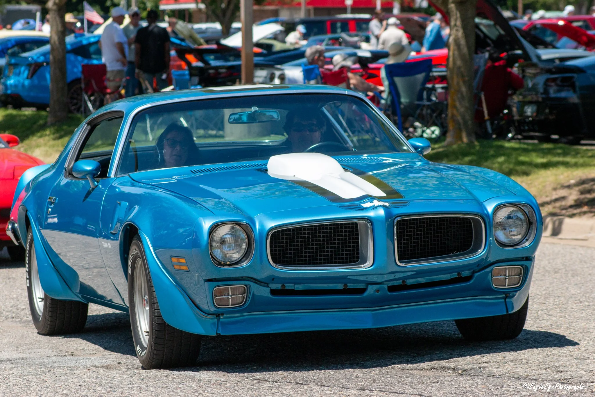 A blue classic muscle car with a white racing stripe on the hood at a car show. The car has a black grille and round headlights, and there are people in the background looking at other cars.