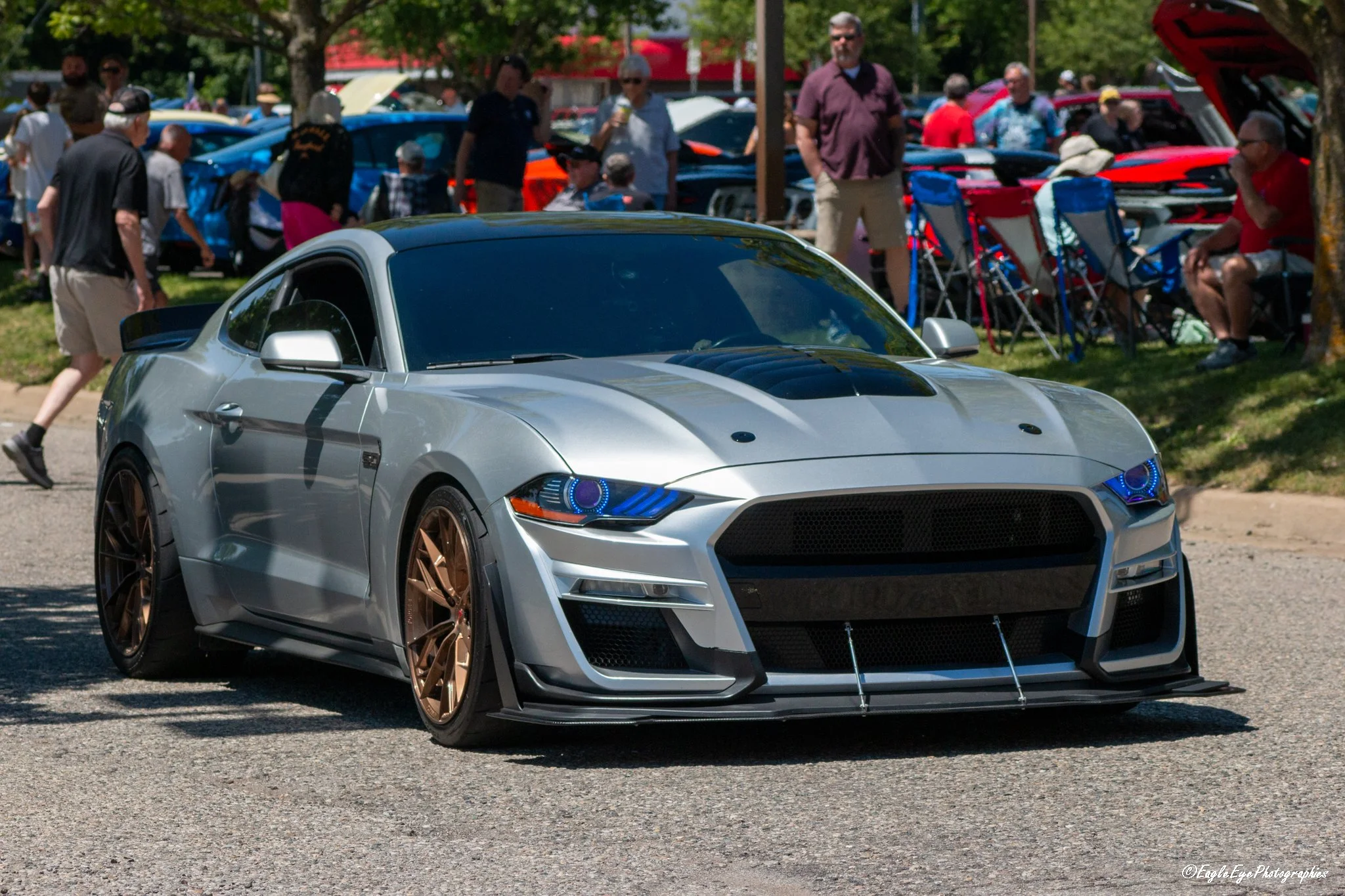 A silver sports car with black and gold rims parked at an outdoor event, with a crowd of people and other cars visible in the background.