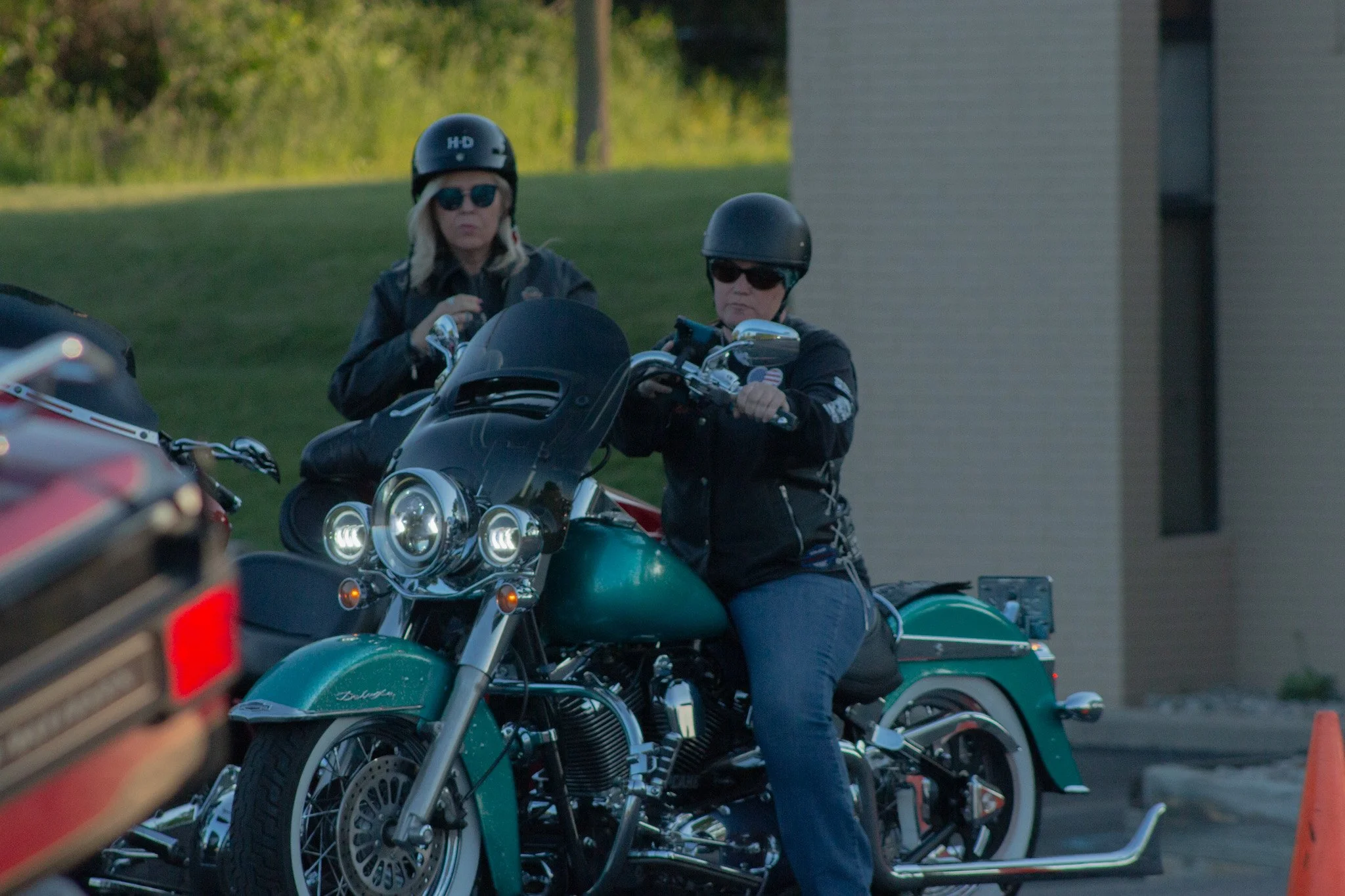 Two women wearing black helmets and sunglasses riding a teal motorcycle during daylight with a person in the foreground and an orange traffic cone beside the motorcycle.