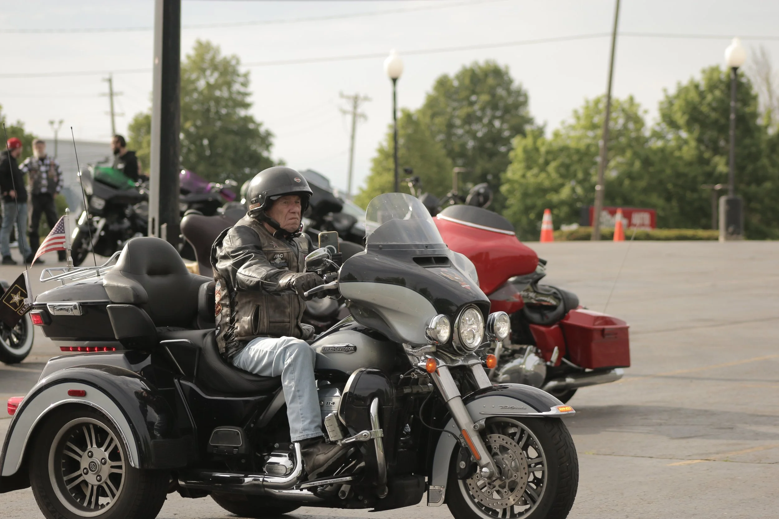 A man wearing a helmet and leather jacket riding a Harley-Davidson trike motorcycle in a parking lot with motorcycles and people in the background.