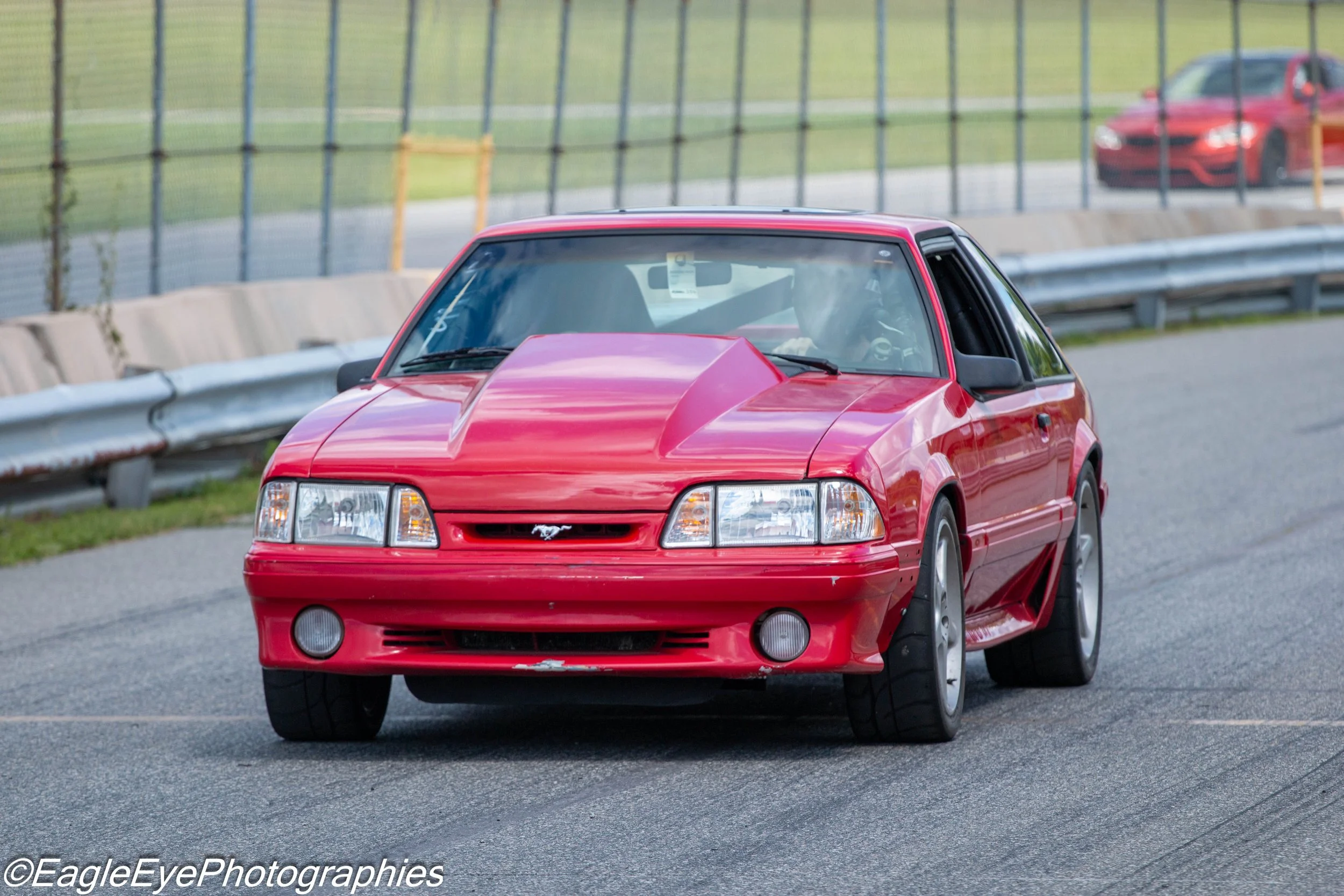 Red Ford Mustang car driving on a paved road with a metal fence on the side and another red car in the background.