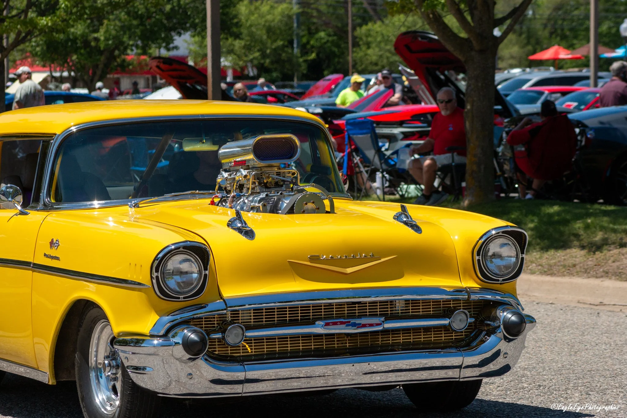 Yellow vintage Chevrolet car with a supercharged engine at a car show, with people and other cars in the background under trees and red umbrellas.