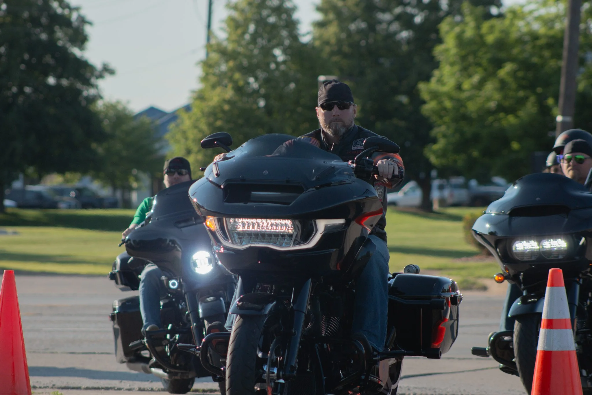 A man with a beard wearing sunglasses, a black cap, and a black vest riding a black motorcycle, with a few other riders on motorcycles riding behind him in an outdoor setting with trees and parked cars in the background.