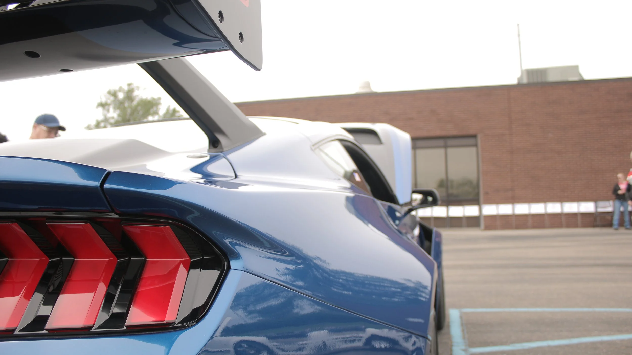 Close-up of the rear of a blue Ford Mustang with a large rear wing, parked near a brick building with glass windows, with a person and a police officer in the background.