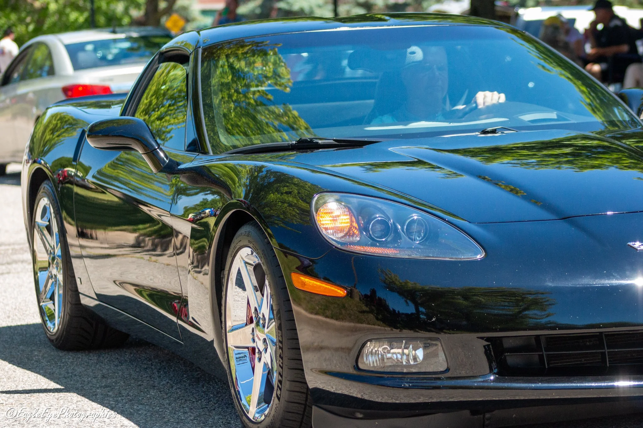 A shiny black sports car parked outdoors, with reflections of green trees on its surface. In the background, there are other parked cars and people at a gathering or event.