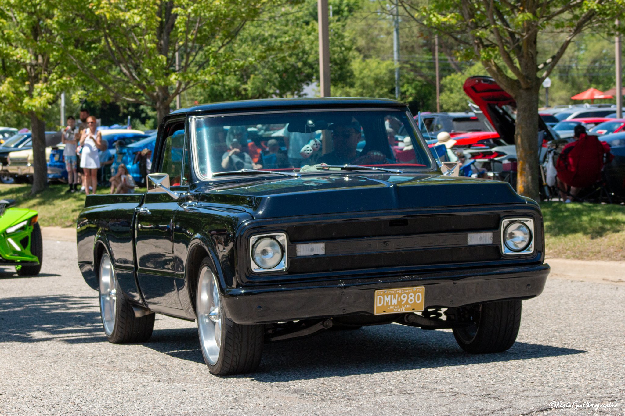 A black vintage car with a Michigan license plate reading DMW 980 parked at an outdoor car show, with other cars and people in the background under trees on a sunny day.