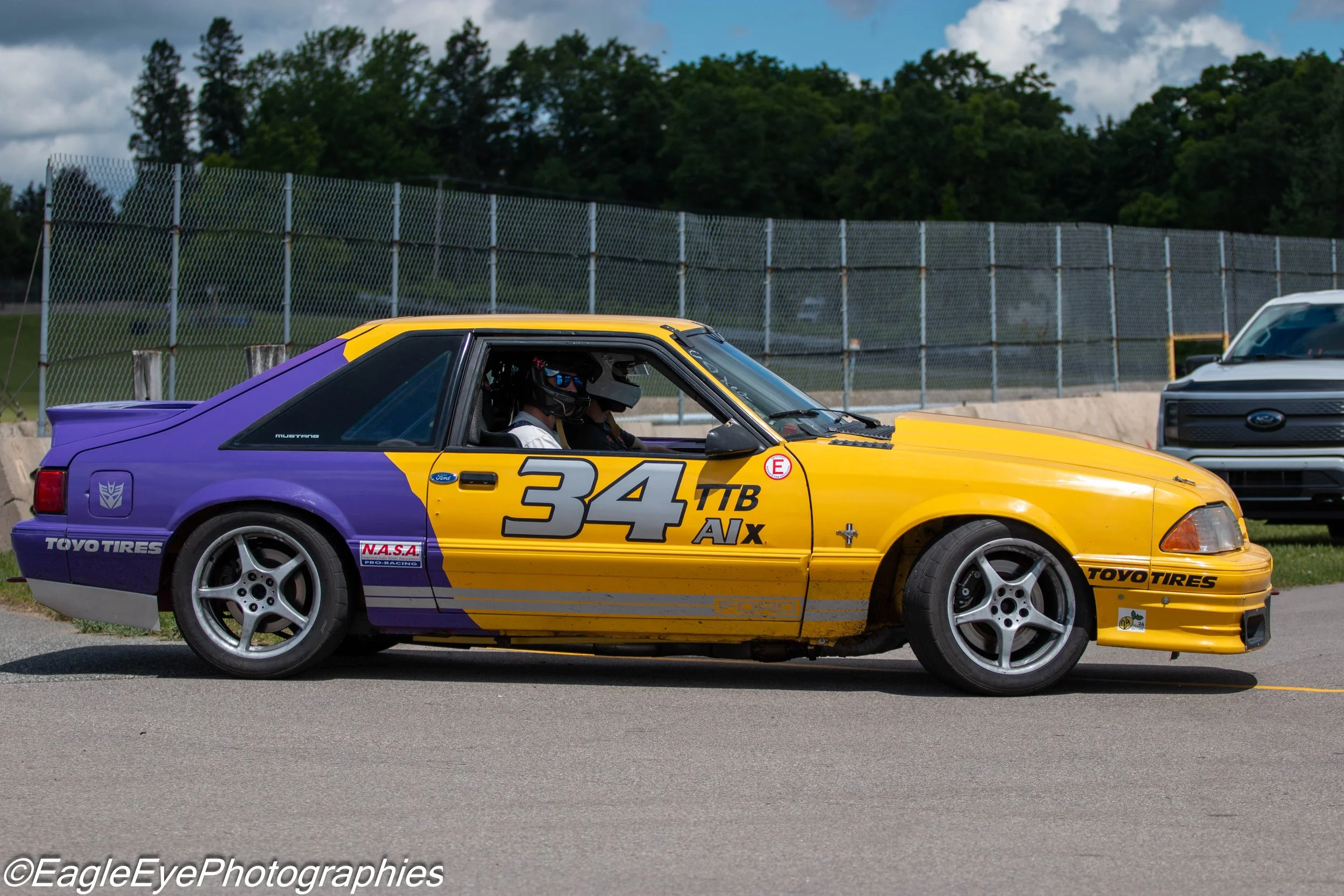 A race car with a purple and yellow paint scheme, the number 34, and various sponsor decals on a racetrack.