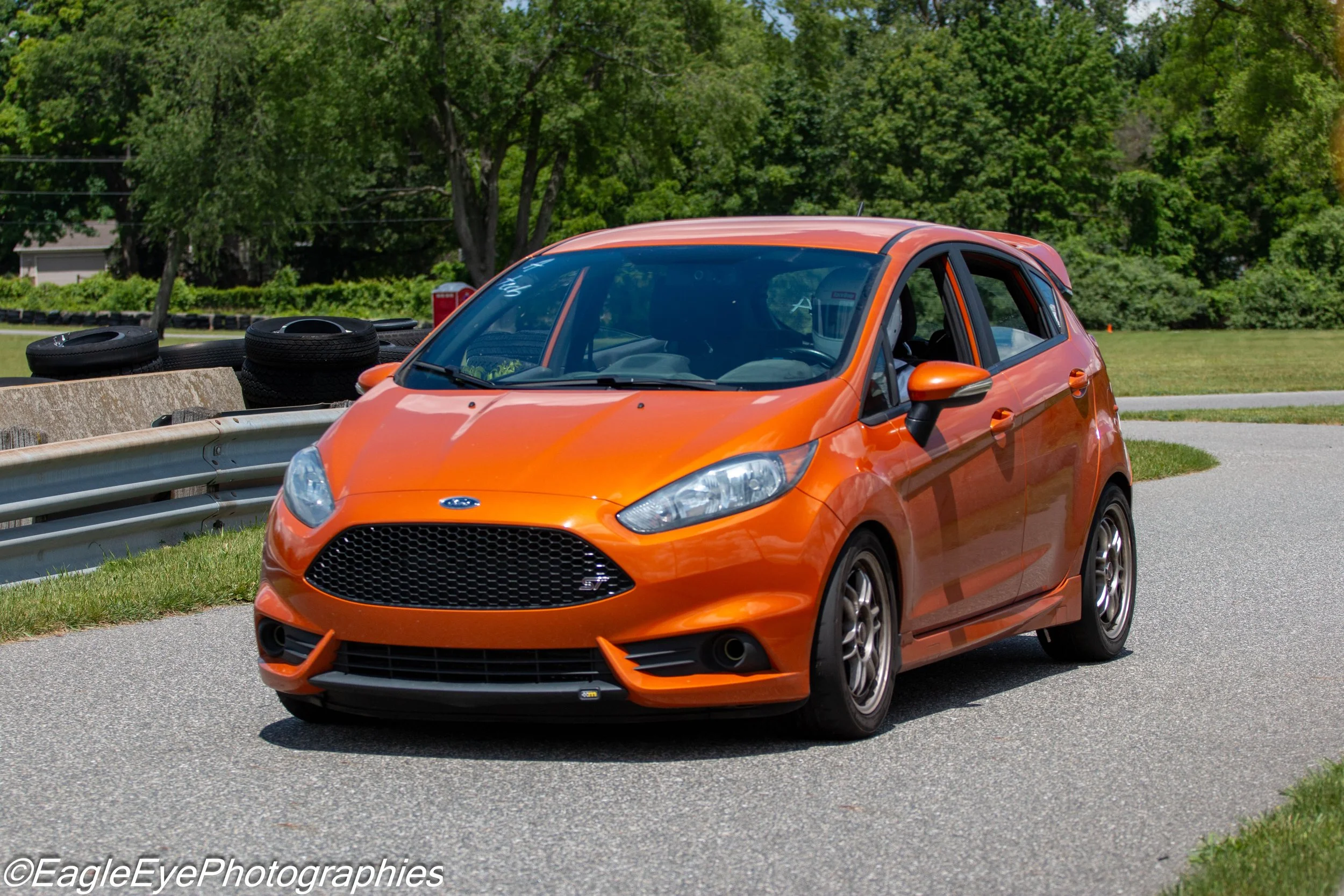 Orange compact car driving on road with tires stacked on roadside and trees in background.