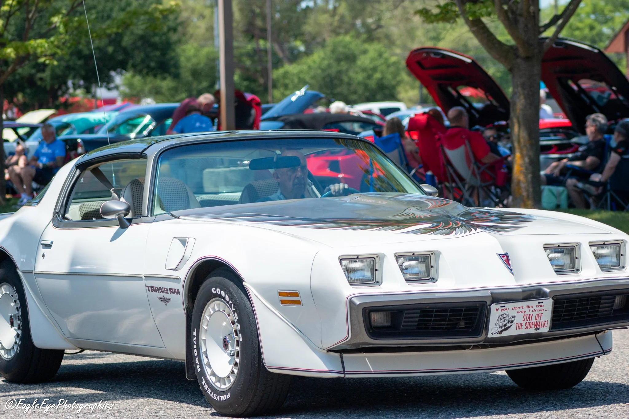 A white Pontiac Trans Am sports car with detailed flames on the hood, parked at an outdoor car show or parade with other classic cars visible in the background. The car has a sign on the front bumper with safety instructions.