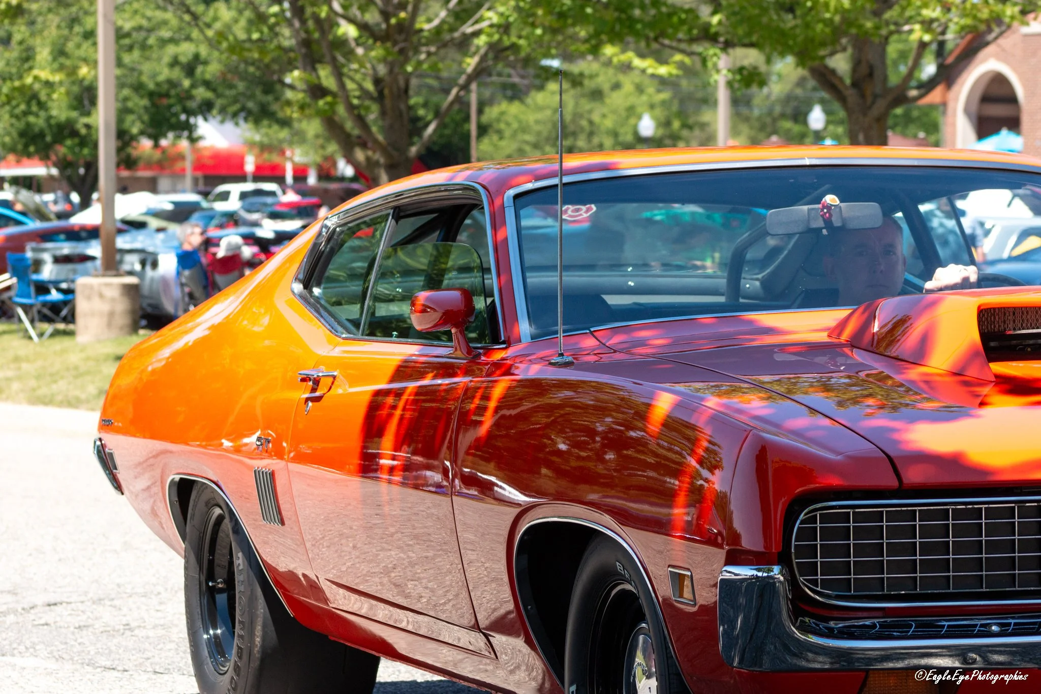 Close-up of a vintage red and orange muscle car at a car show, with a crowd and trees in the background.
