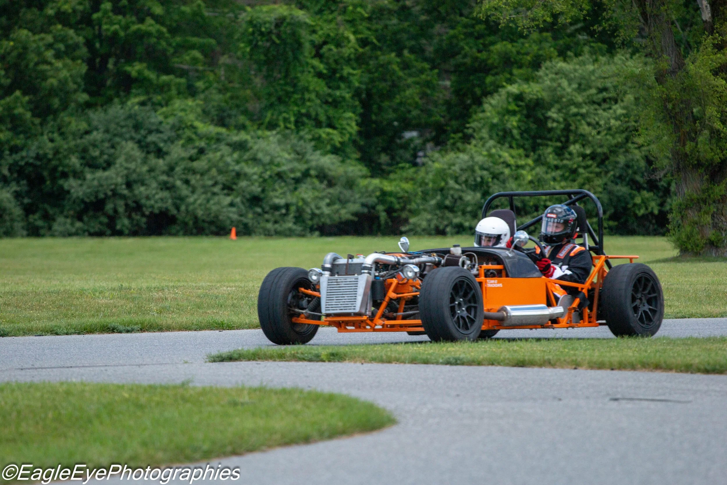 A person wearing a helmet sitting in an orange open-wheel racing car on a grassy area with trees in the background.