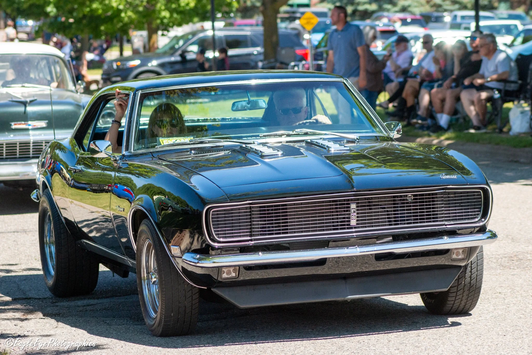 A black vintage Chevrolet Camaro car at an outdoor car show, with people sitting on benches and standing in the background, under trees on a sunny day.