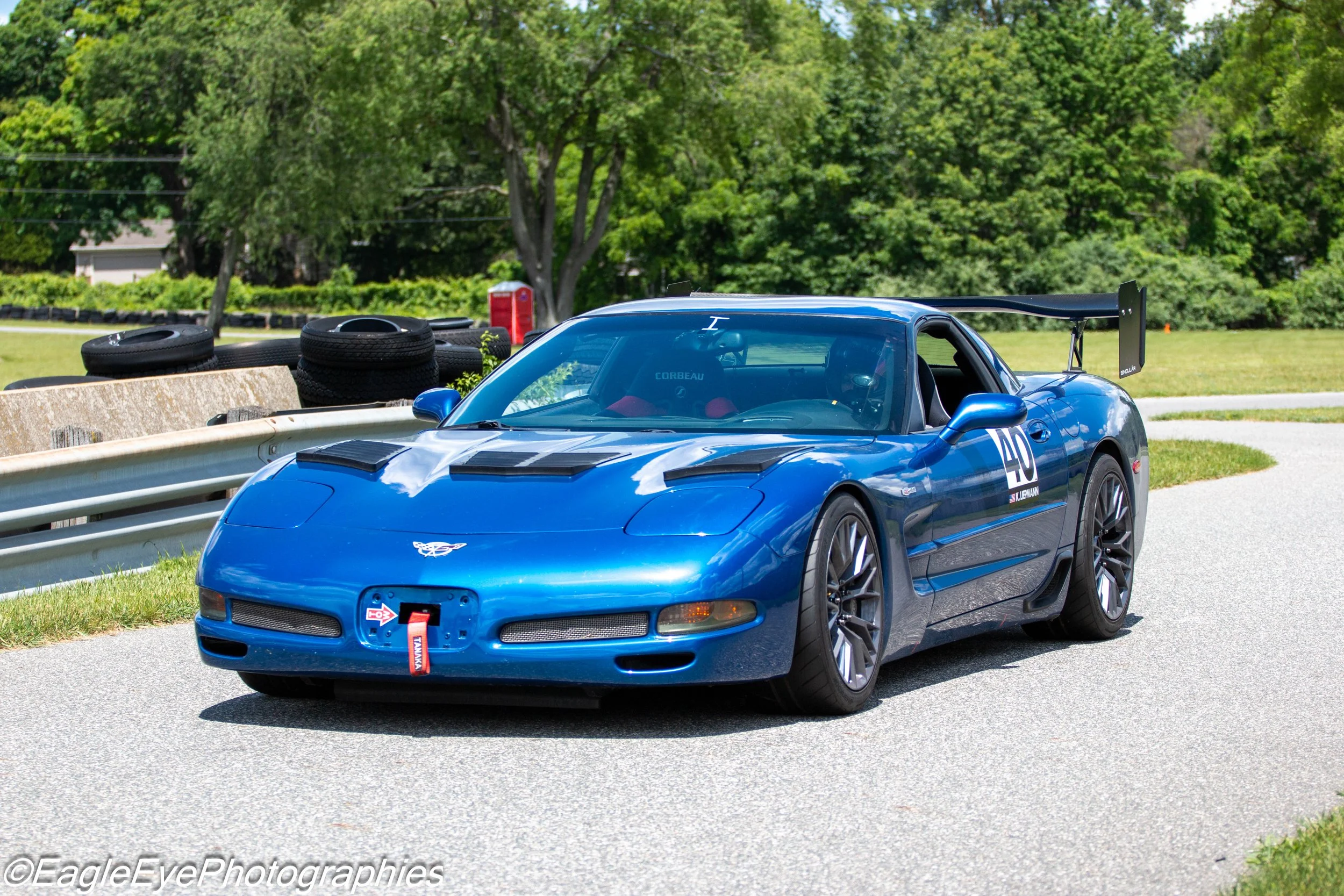 A blue Chevrolet Corvette race car with the number 40 on the side, parked on a road near a grassy area with tires stacked behind it and a forested background.