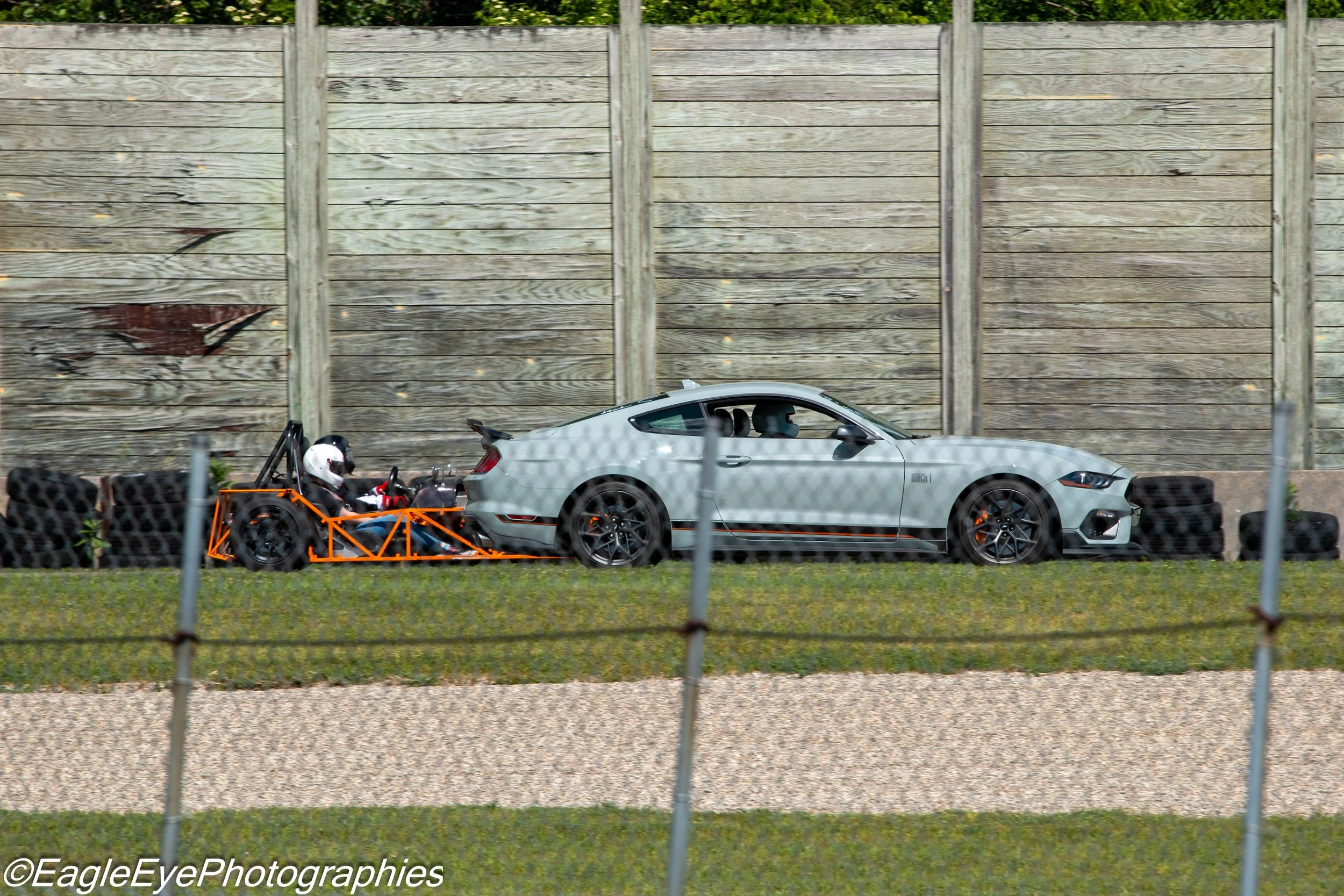 A silver sports car on a race track with a go-kart driver in front of it and a wooden wall in the background.