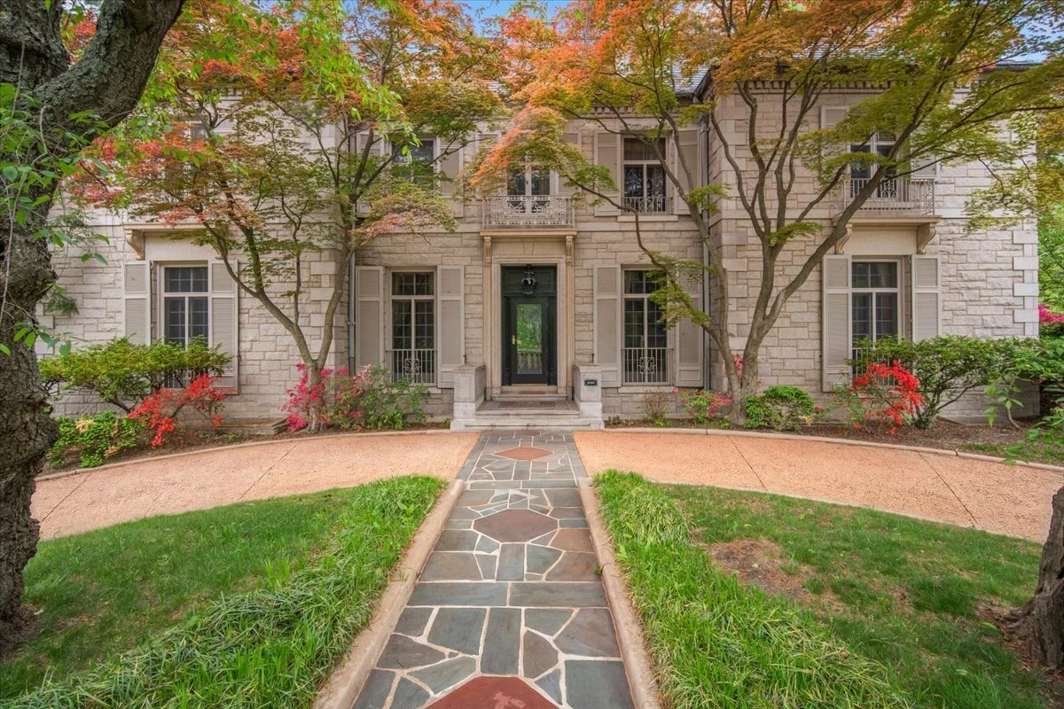 Front view of a stone house with a black door, surrounded by trees and colorful shrubs, with a pathway leading to the entrance.
