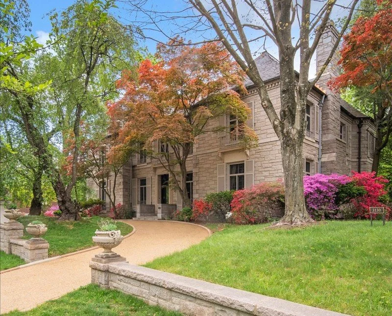 A historic stone mansion surrounded by lush greenery and blooming flowers, with a gravel pathway leading up to the front door.