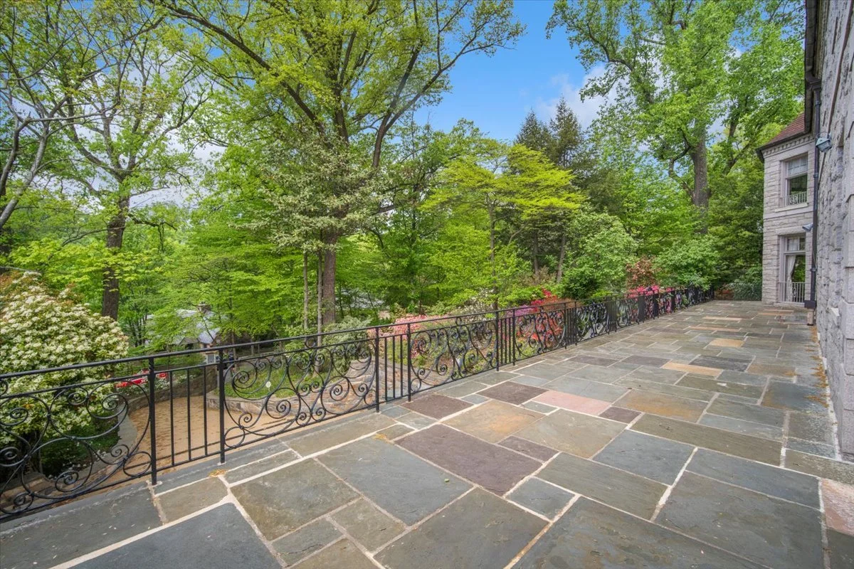 Stone patio overlooking lush green trees, surrounded by a decorative black wrought iron railing, with a stone house wall on the right.