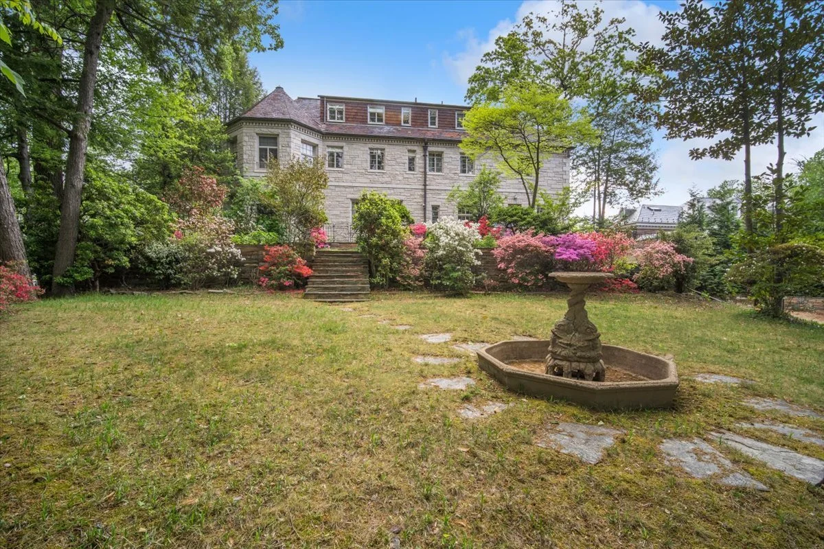 A spacious backyard with a grassy lawn, stone steps leading up to a large stone house, and a vintage stone fountain in the yard. The landscape is lush with flowering bushes and trees under a clear blue sky.