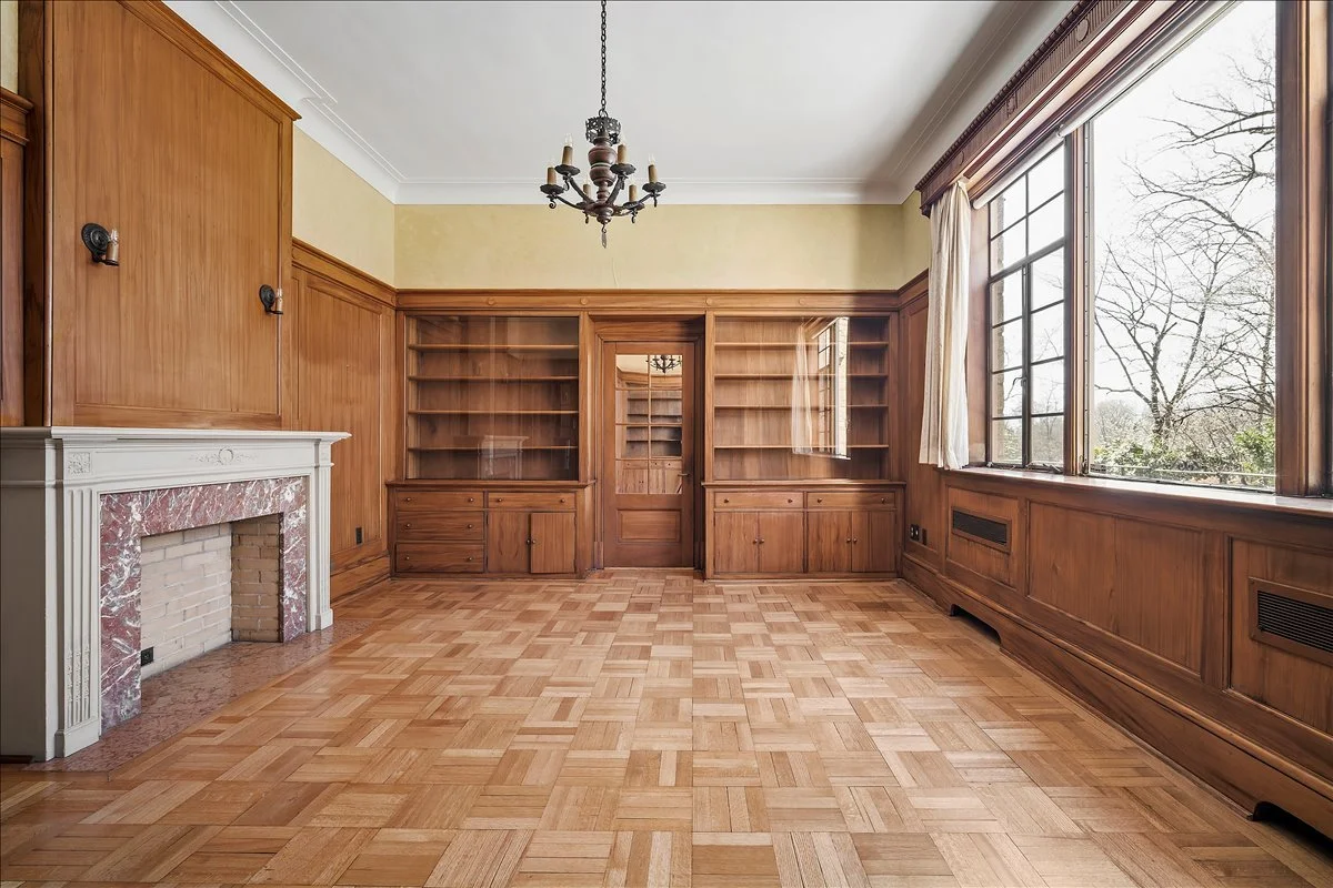 Empty living room with parquet flooring, wood-paneled walls, large windows with curtains, built-in wooden bookshelves, a fireplace with marble trim, and a chandelier hanging from the ceiling.