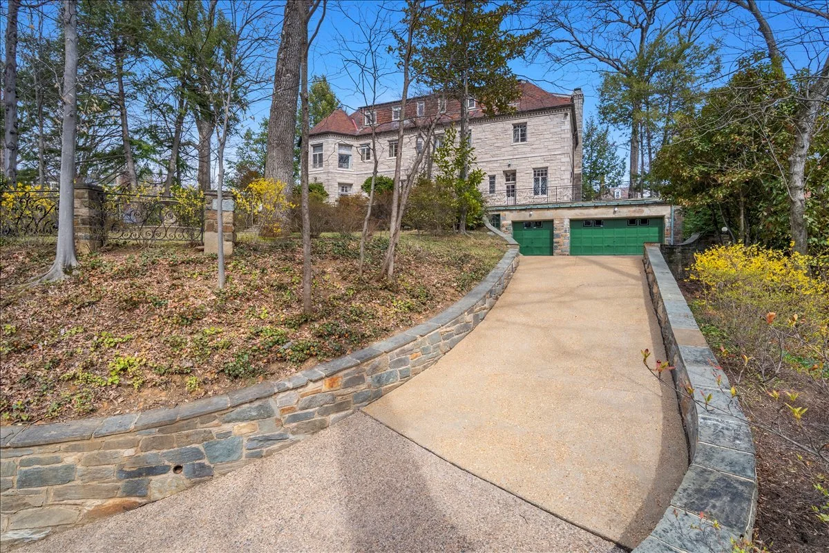 A house with a stone exterior and a red roof, seen from the bottom of a sloped driveway lined with a stone retaining wall, surrounded by trees and shrubs.