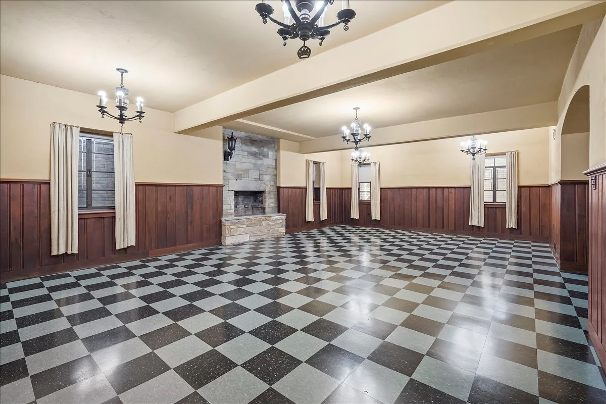 Empty living room with checkered tile flooring, wooden wall paneling, stone fireplace, multiple windows with cream curtains, and black chandeliers on the ceiling.