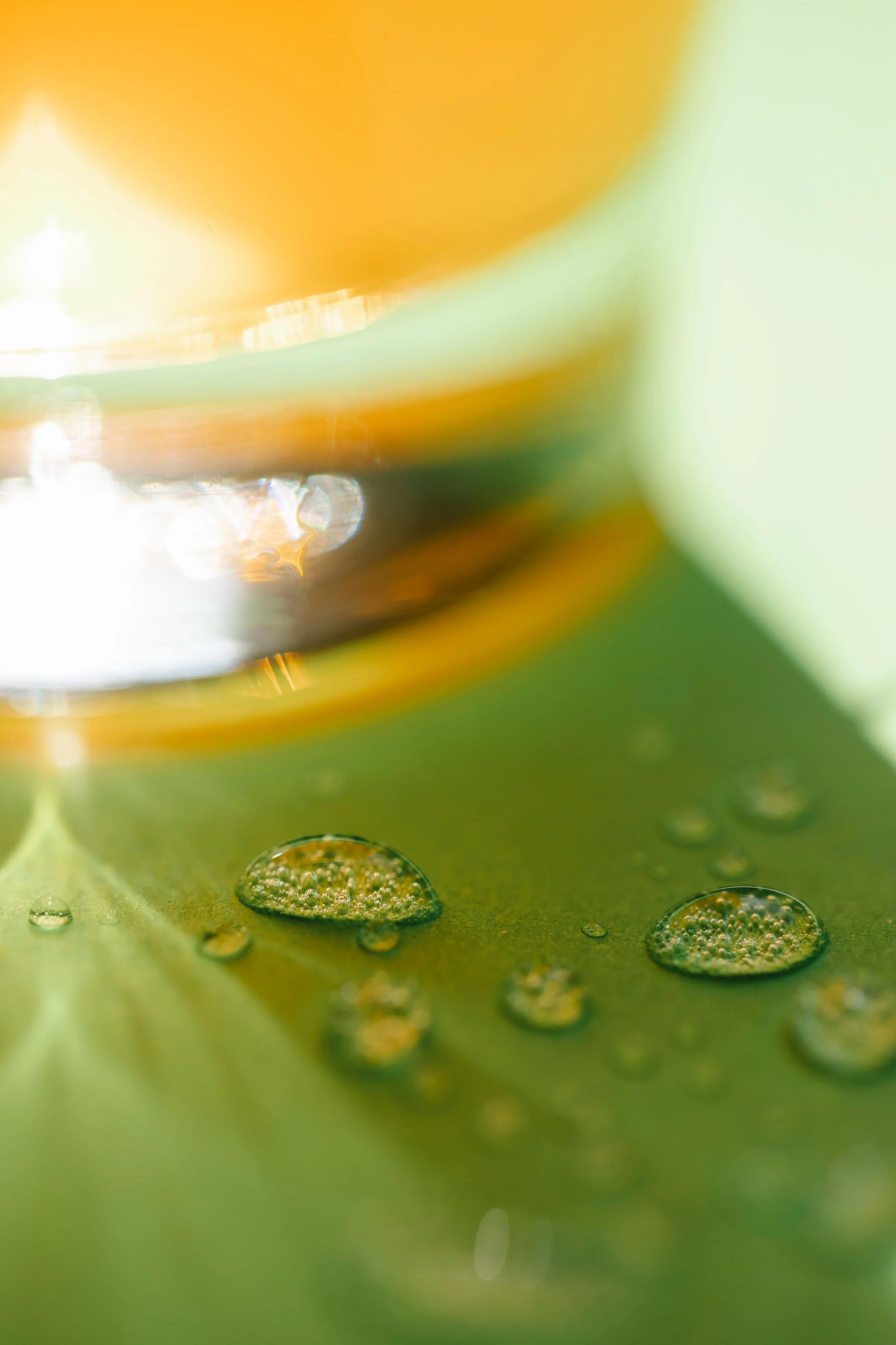 Close-up view of water droplets on a green leaf with a blurred yellow object in the background.