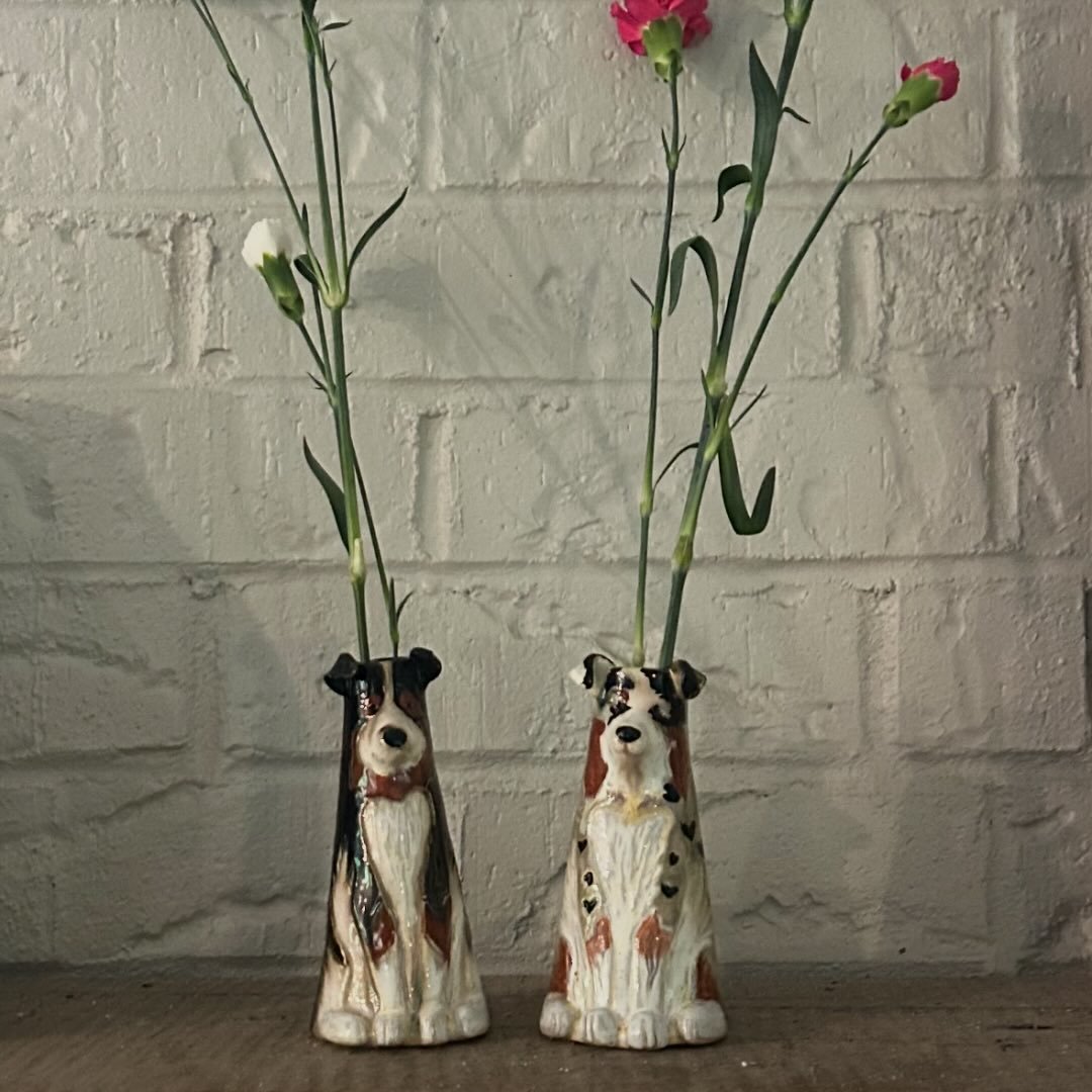 Two handmade ceramic dog-shaped vases with flowers on a wooden surface against a white brick wall.