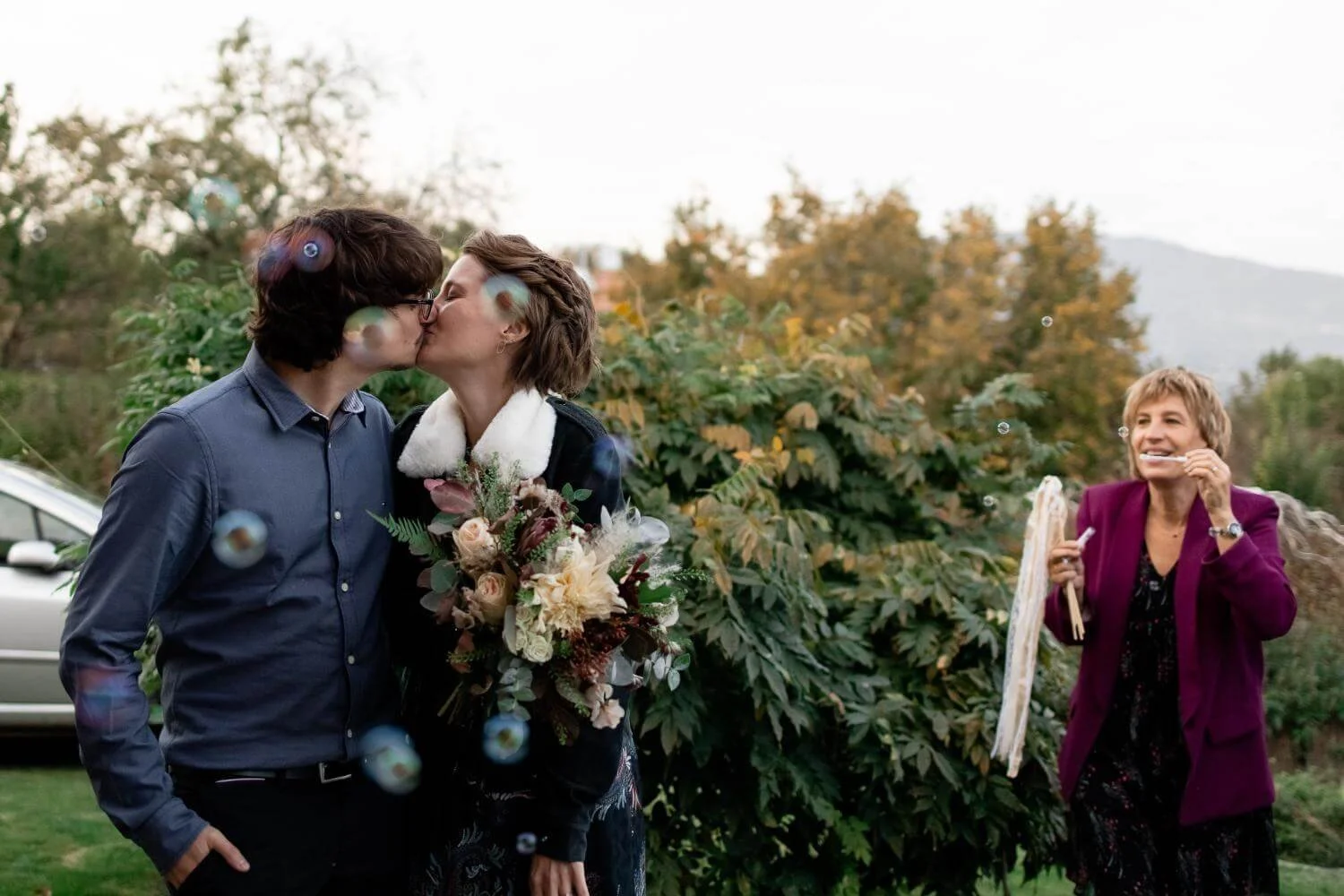 A couple kisses outdoors during daytime, with a woman in a purple blazer smiling and holding a napkin and a face mask nearby, amid greenery and autumn trees.