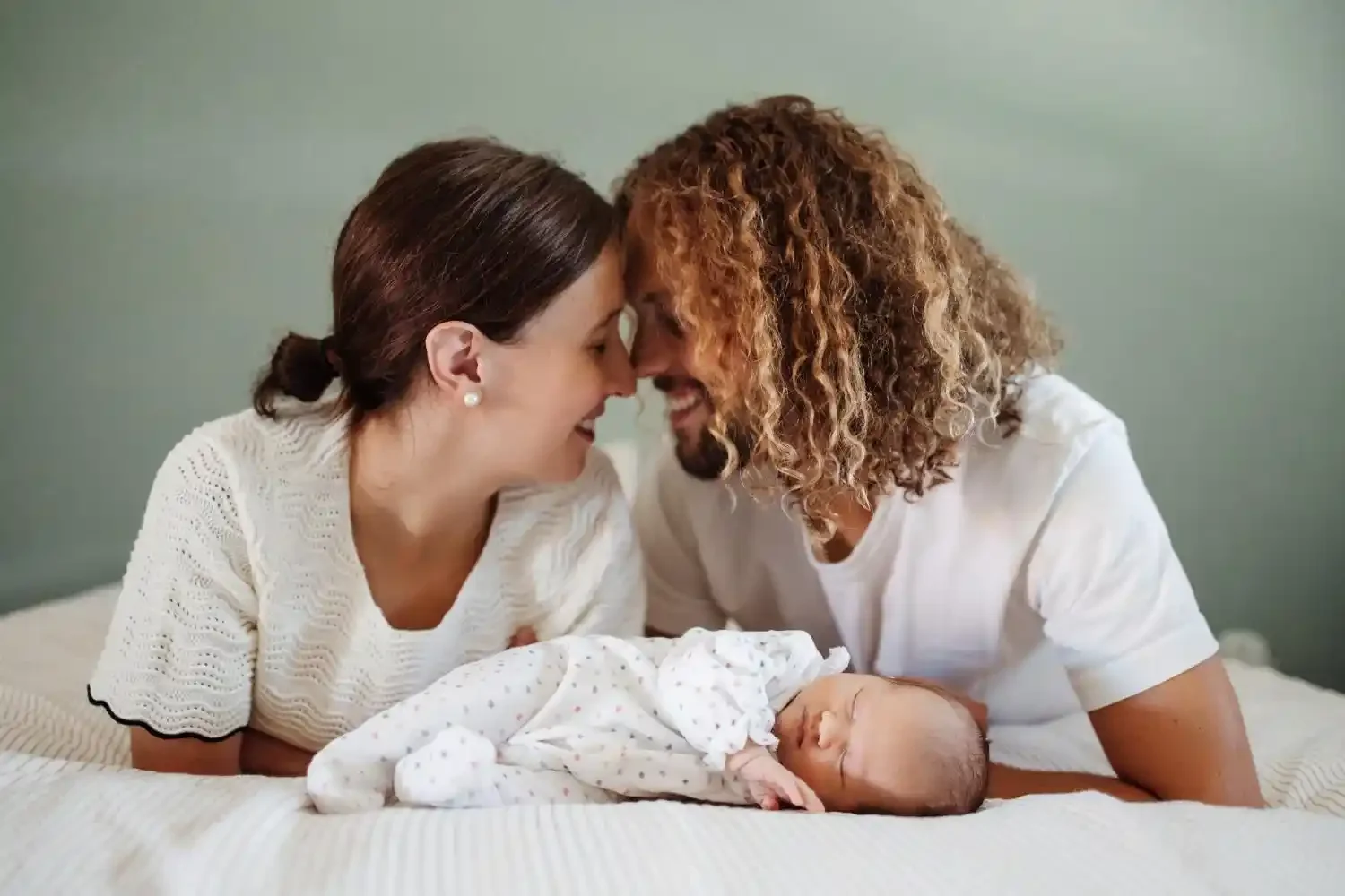 Parents front contre front, souriants, autour de leur nouveau-né endormi sur le lit, séance photo naissance à domicile à Villeneuve-d’Ascq