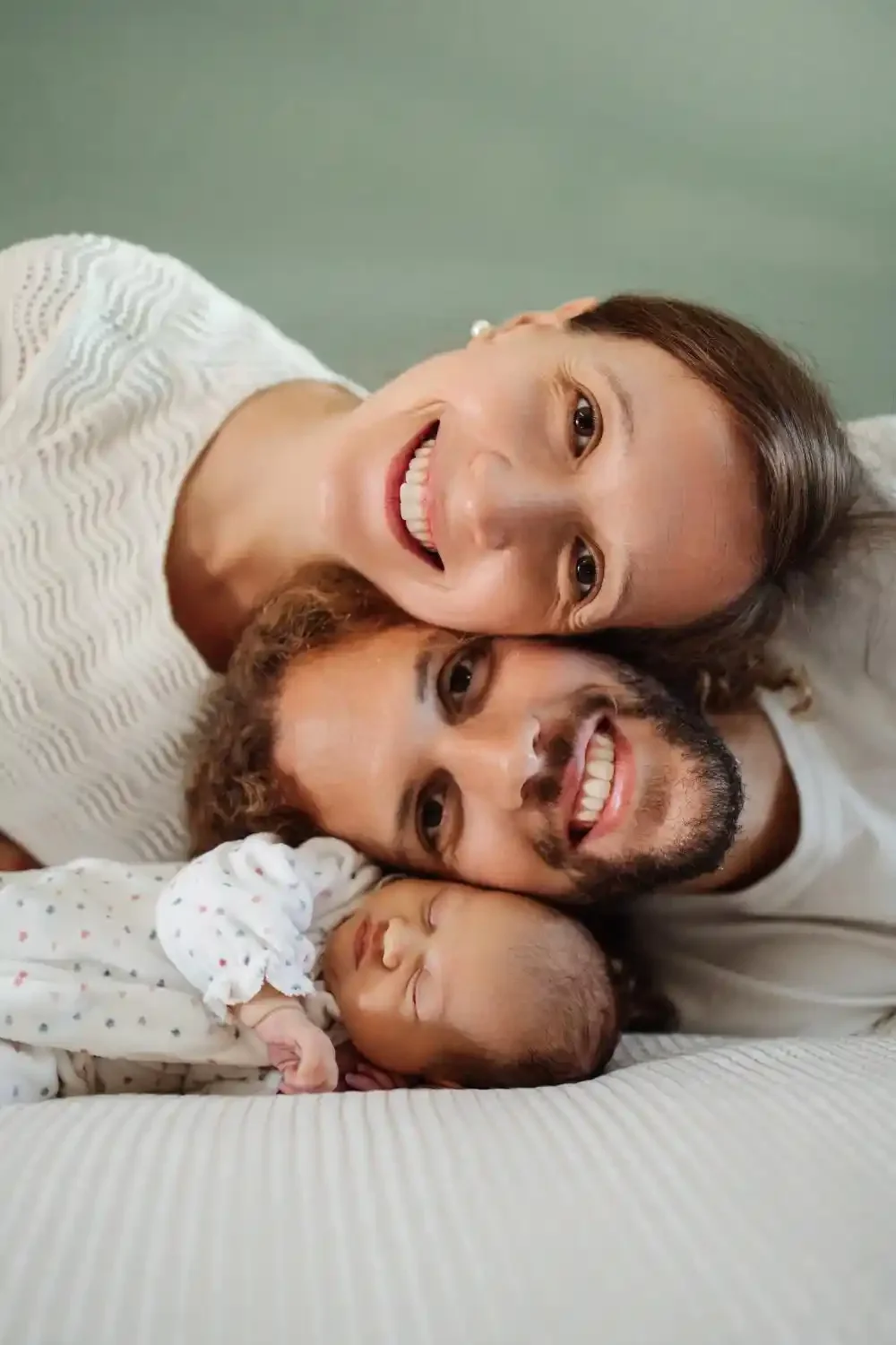 Parents joue contre joue et sourire aux lèvres, leur nouveau-né allongé devant eux, séance famille à domicile