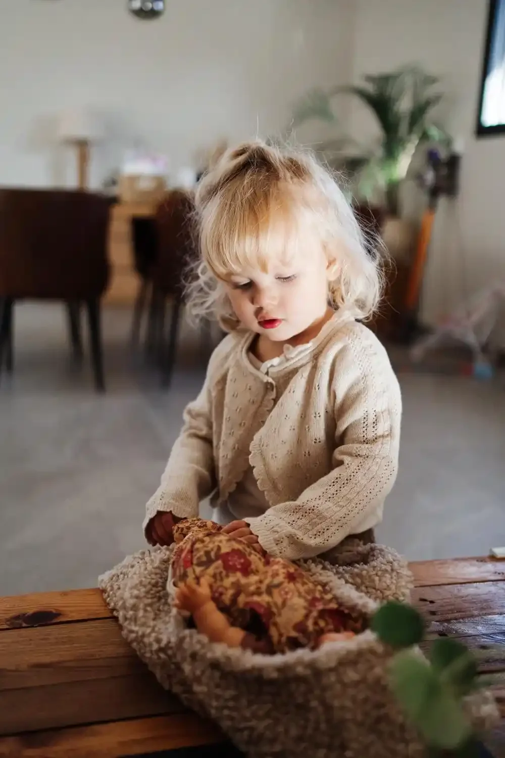 Grande sœur de 2 ans concentrée sur sa poupée installée dans un panier, photo lifestyle d’enfant à la maison