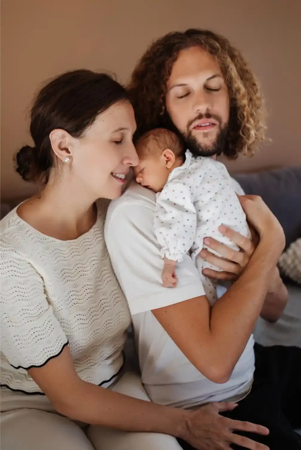 Parents joue contre joue autour de leur nouveau-né, les yeux fermés, sur le canapé familial
