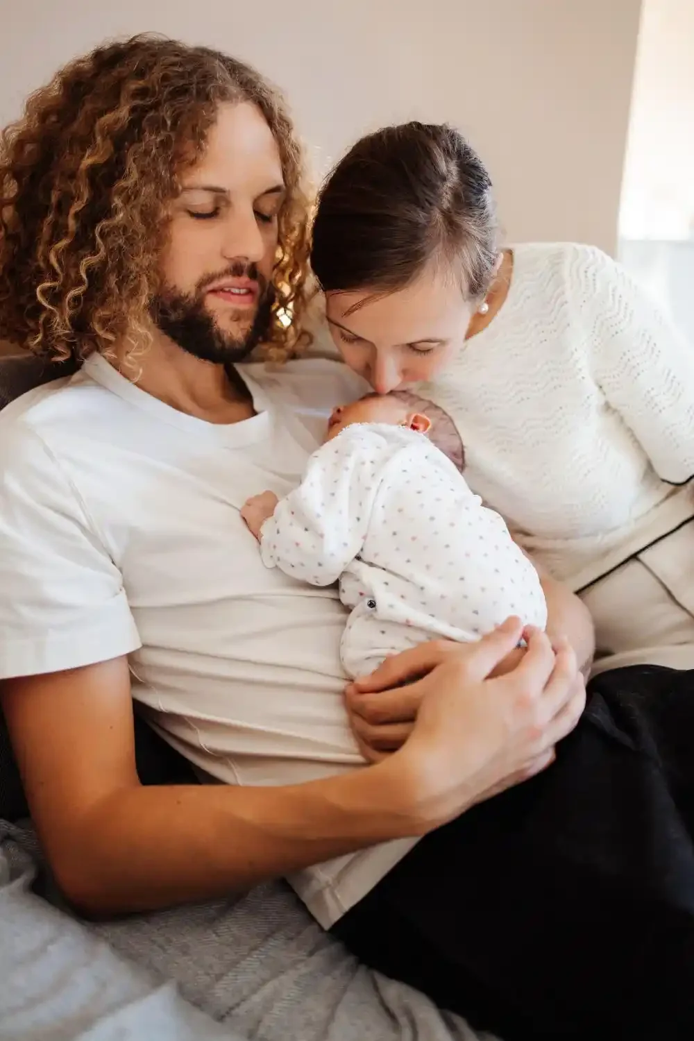 Papa embrasse la tête de son nouveau-né pendant que maman les regarde, photo lifestyle de naissance