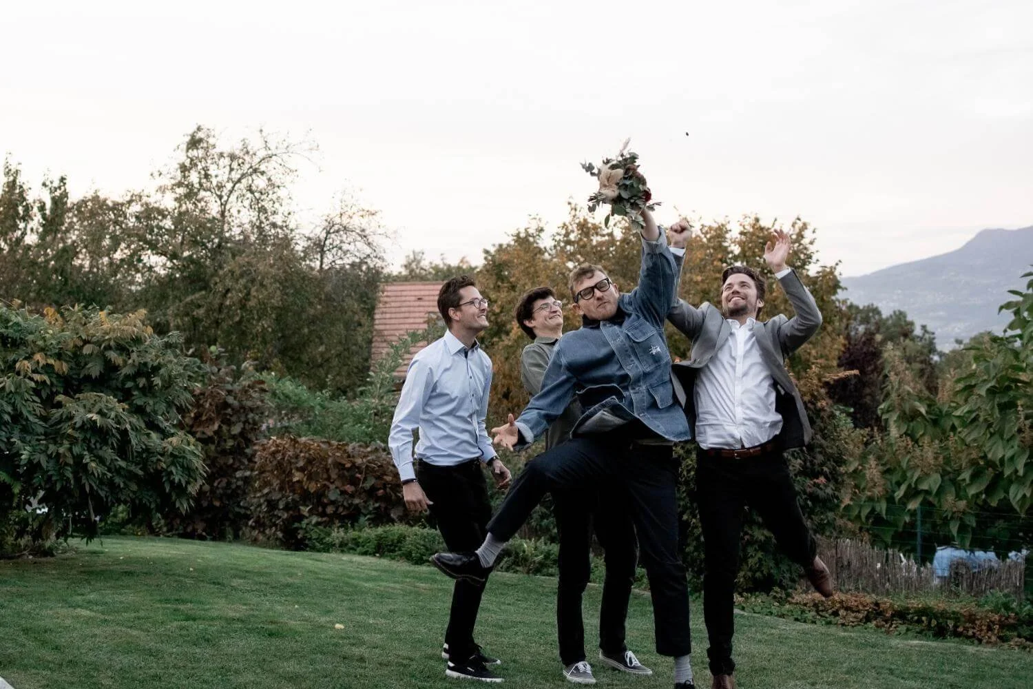Group of five people, four men and one woman, outdoors on a lawn, celebrating a wedding or special occasion, with one man holding a bouquet of flowers high in the air and others smiling and cheering.