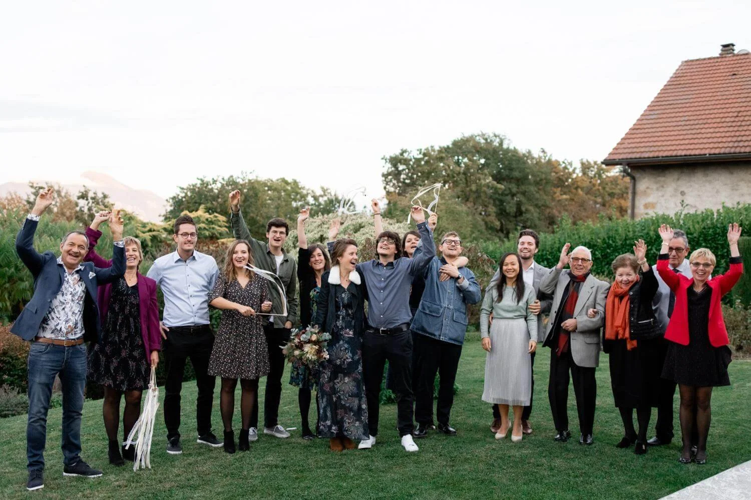 Group of people celebrating outdoors, with friends and family, in a garden, holding balloons and ribbons, smiling, and raising their hands.