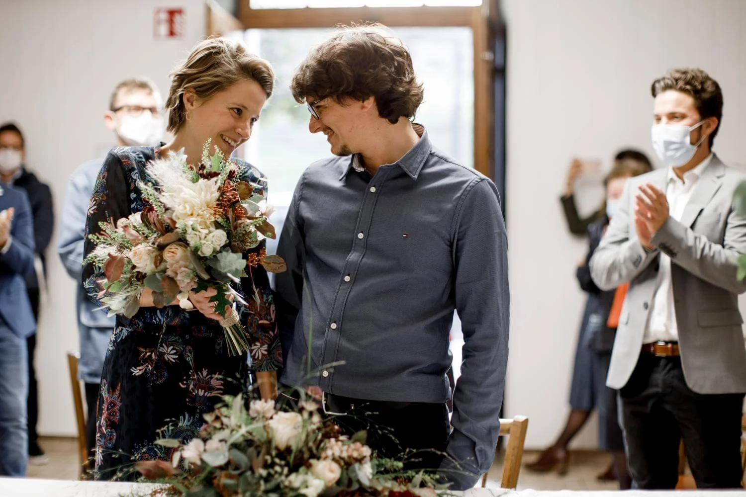 A woman holding a bouquet of flowers is smiling at a man in a blue shirt at a gathering. People in the background are wearing masks and clapping.