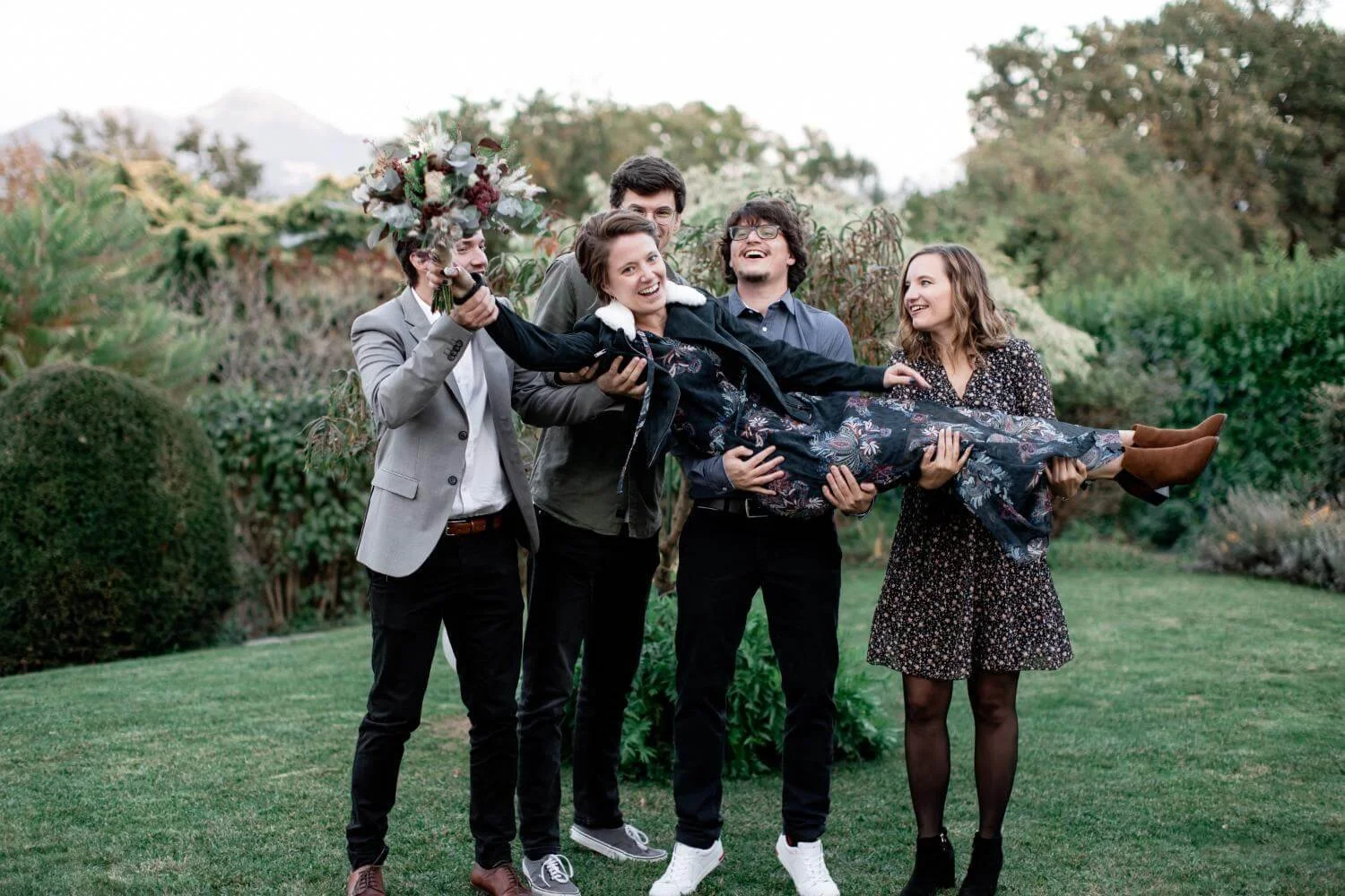 Group of five young adults outdoors, with the man on the left holding a woman in a floral dress, who is smiling and holding a bouquet of flowers, while others around are laughing and supporting her in a playful pose."