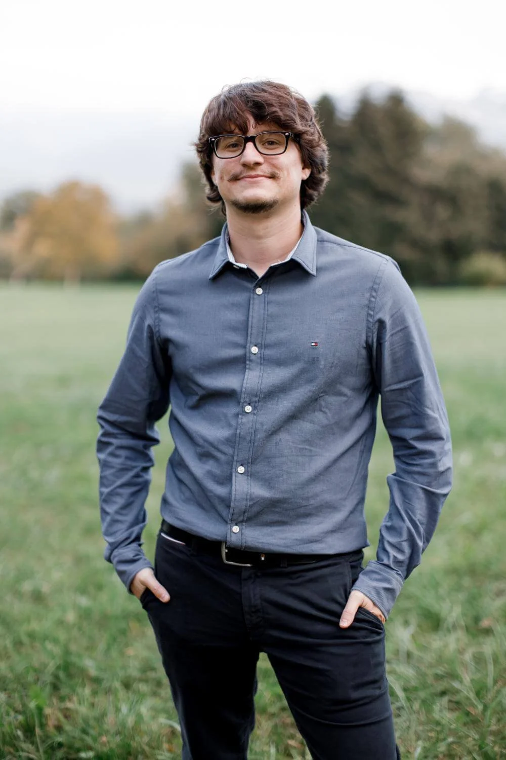 A young man with glasses, wearing a blue button-up shirt and black pants, standing outdoors in a grassy field with trees in the background, smiling at the camera.