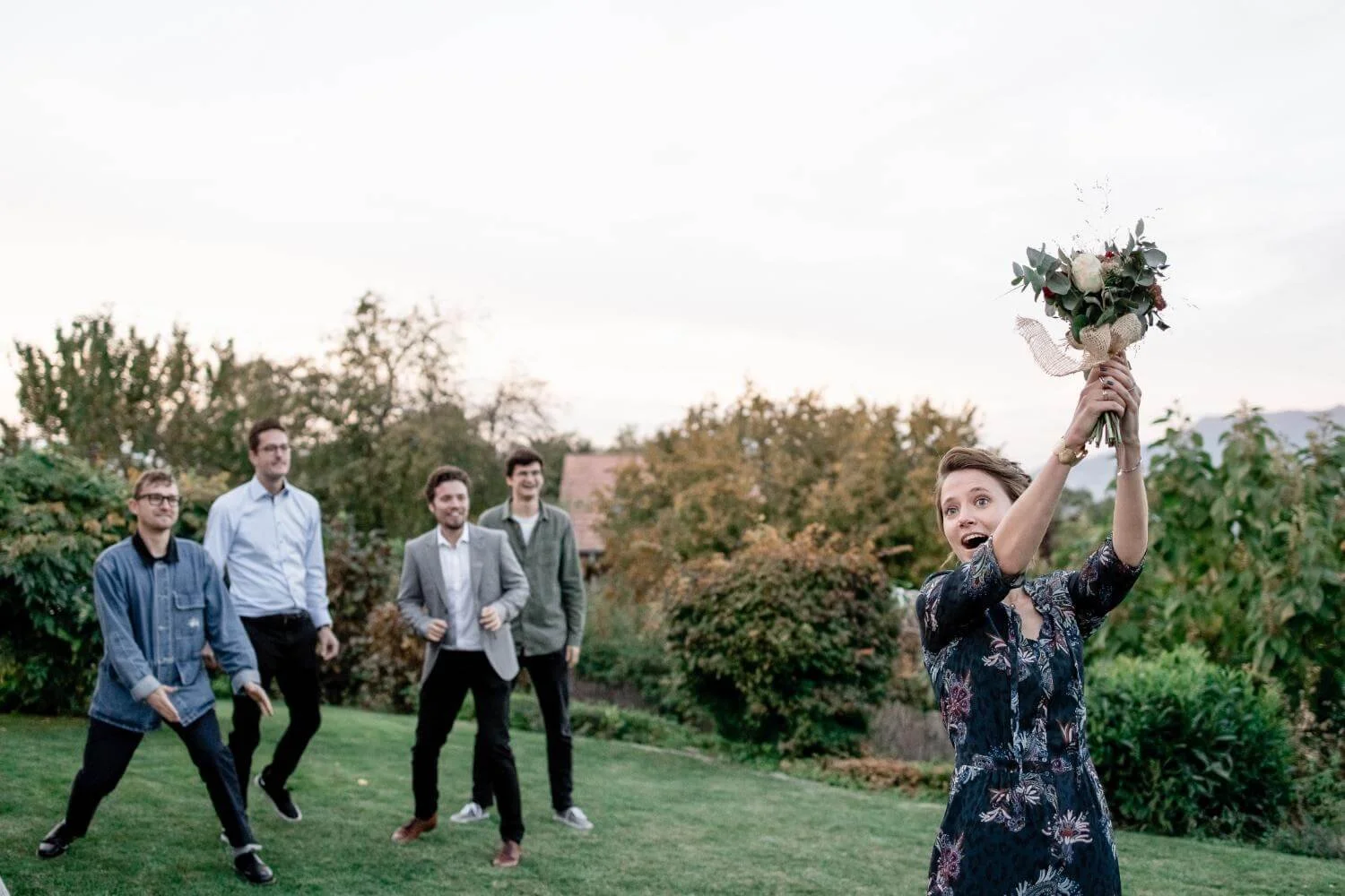 A woman in a floral dress is holding a bouquet of flowers above her head with excitement, while four men stand in the background on a grassy area with trees and bushes, smiling and watching her.