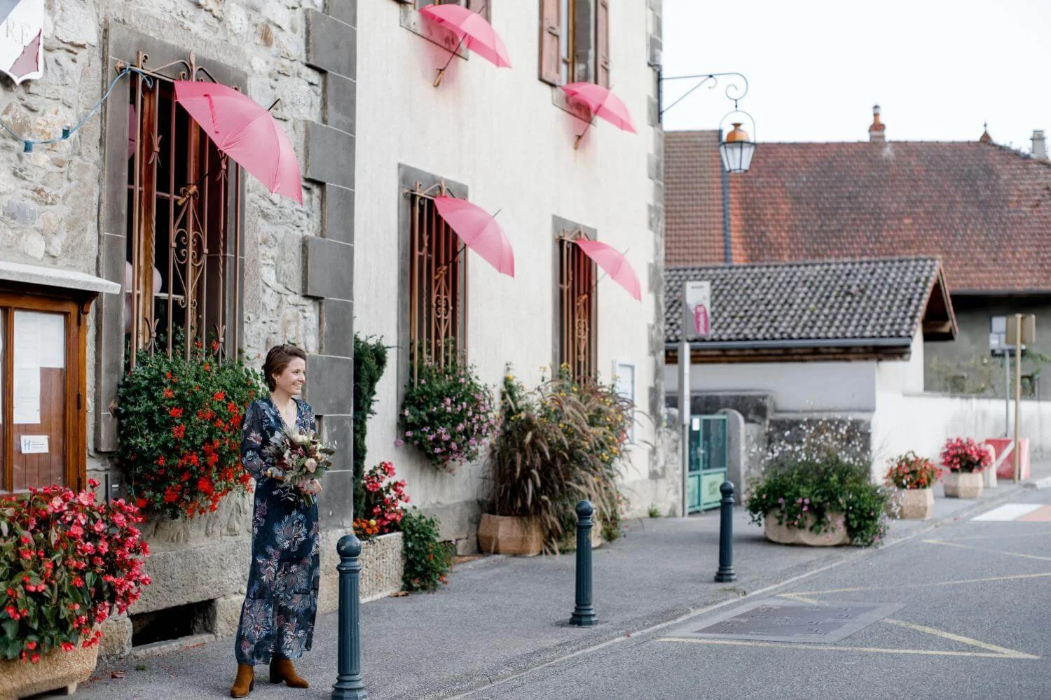A woman in a dark floral dress standing on a sidewalk holding a bouquet of flowers, with pink umbrellas attached to windowsill planters above her, in a quaint street scene with stone and stucco buildings and colorful potted plants.