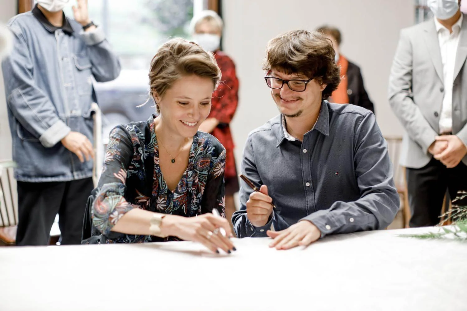 A woman and a man sitting at a table, smiling and signing a document, with several people standing around them in a bright room.