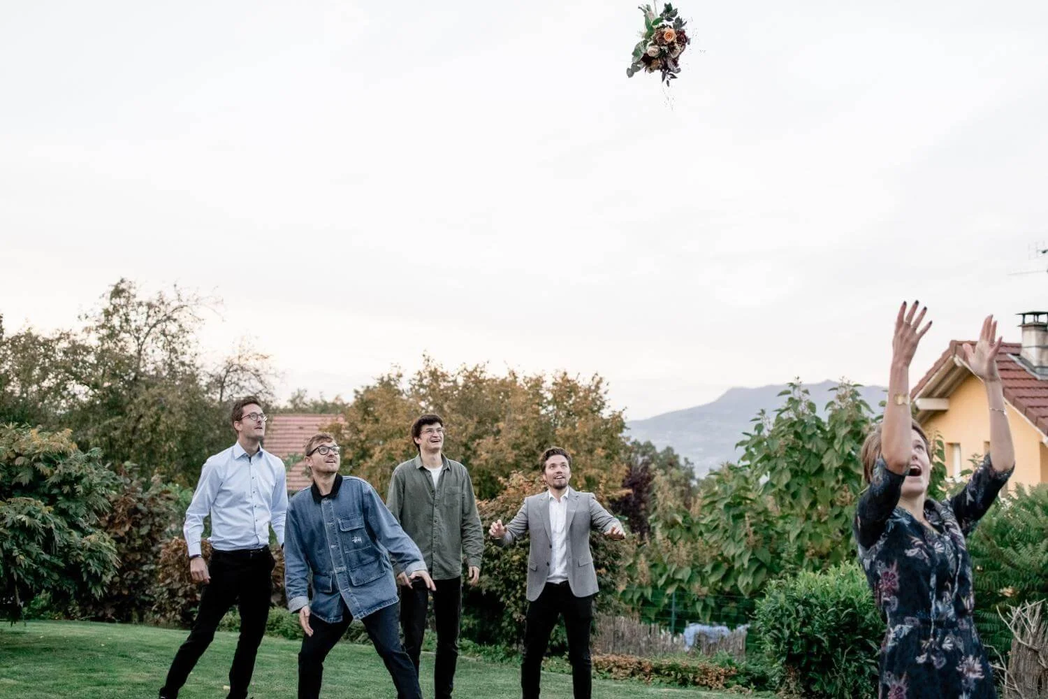 A woman in a dark floral dress throws a bouquet into the air as five men in casual and semi-formal attire watch and smile in a garden with trees, bushes, a house, and mountains in the background.