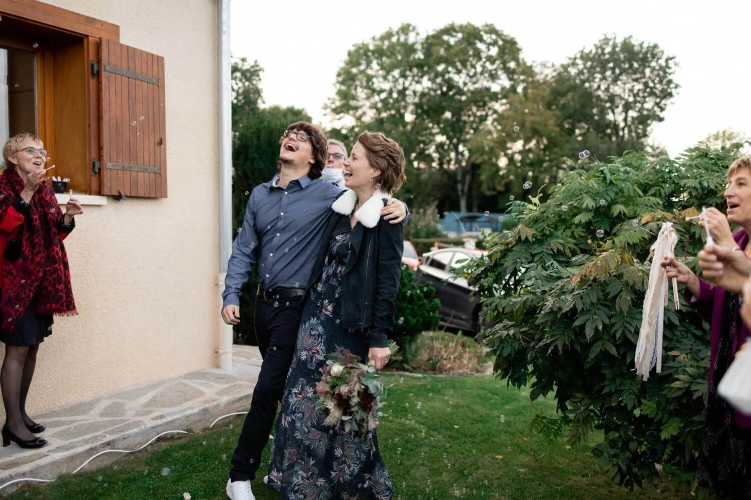 A celebration outside a house with people blowing bubbles and smiling, including a woman with a bouquet of flowers hugging a man, and others enjoying the moment.
