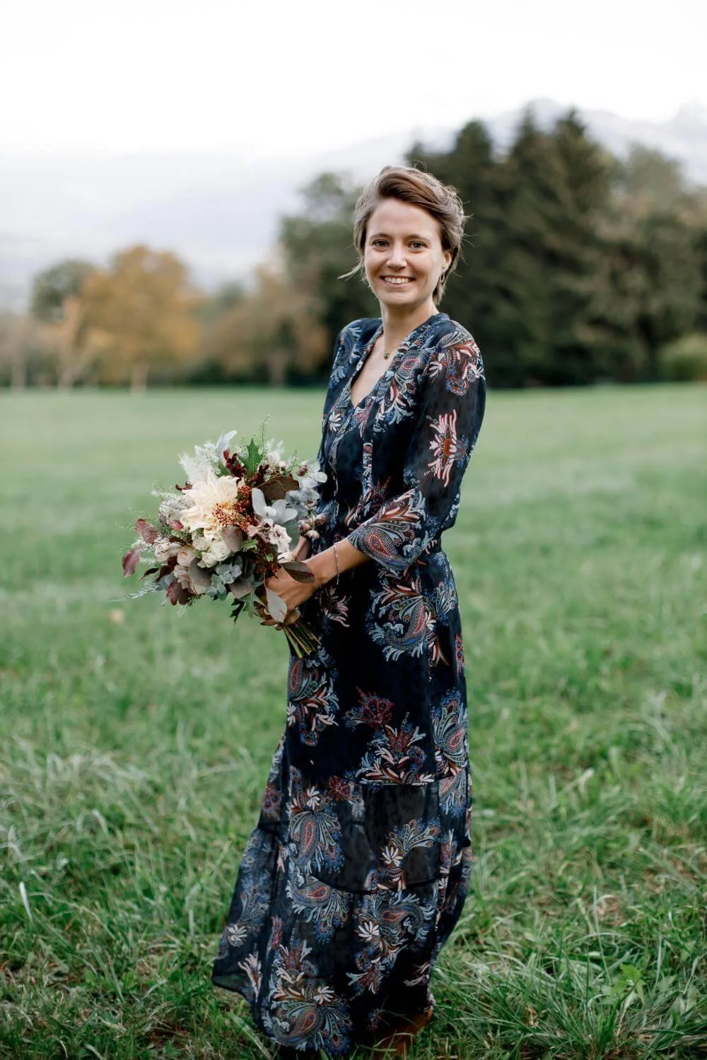 A woman in a dark, floral patterned dress standing on a grassy field, holding a bouquet of flowers, smiling at the camera with trees and a cloudy sky in the background.