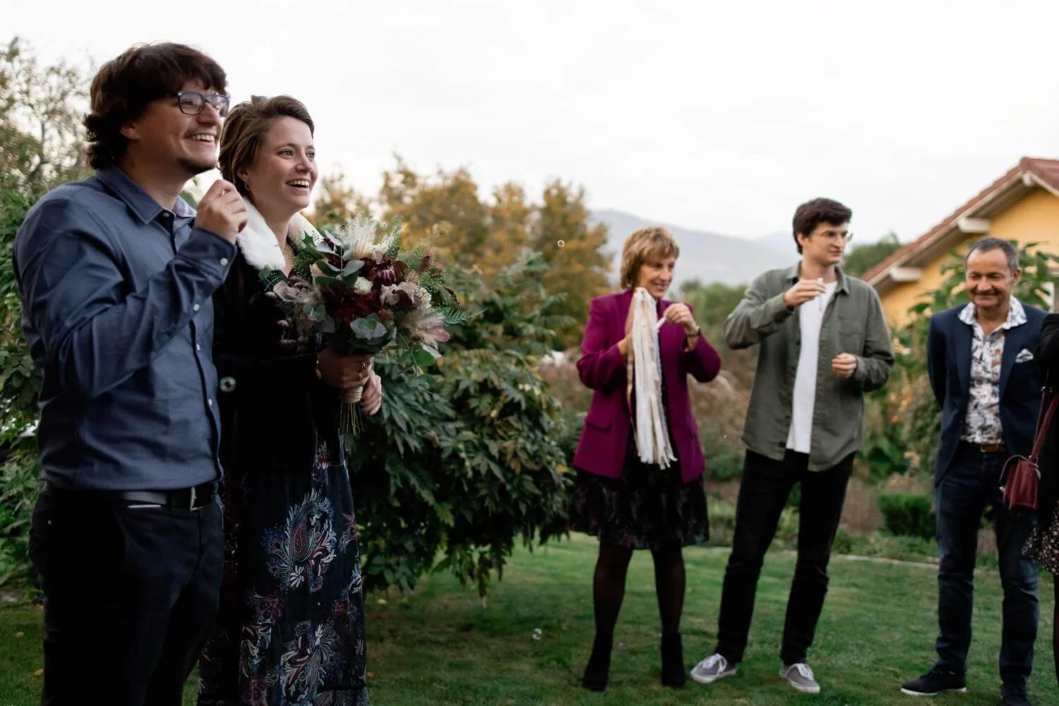 Group of five people standing outdoors, smiling, with trees and a house in the background. A woman in the center holds a bouquet of flowers.